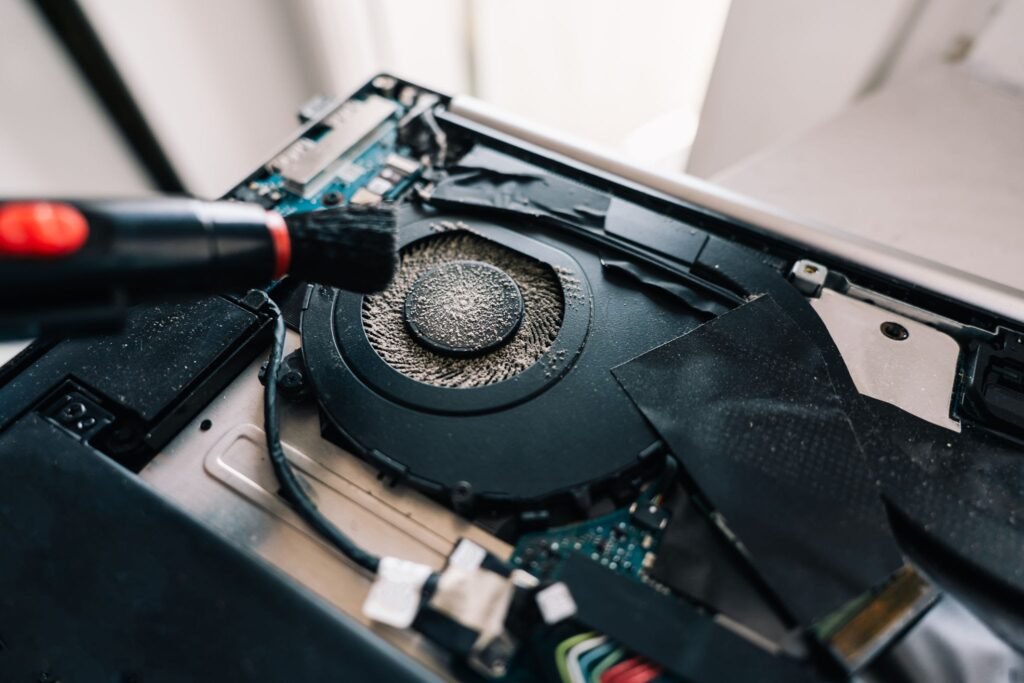 Hands using a small brush and vacuum to carefully clean thick dust out of an exposed laptop cooling fan.