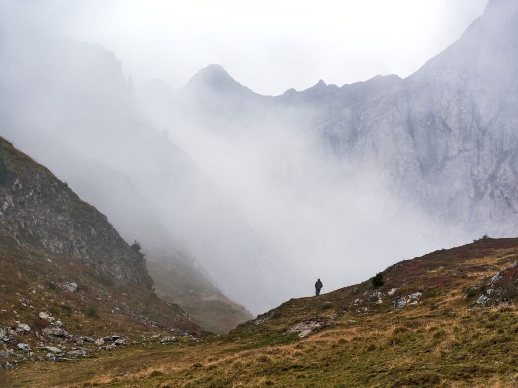 A lone hiker walking down a steep, grassy hillside shrouded in thick, heavy white fog.