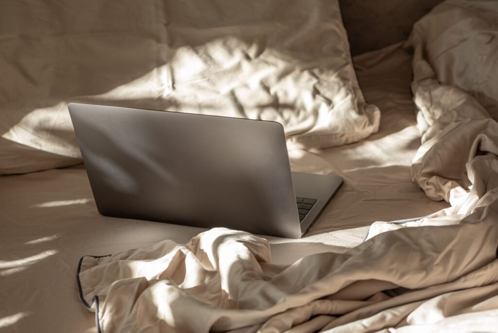 A modern silver laptop resting on a soft, messy bed surrounded by thick blankets in the sunlight.